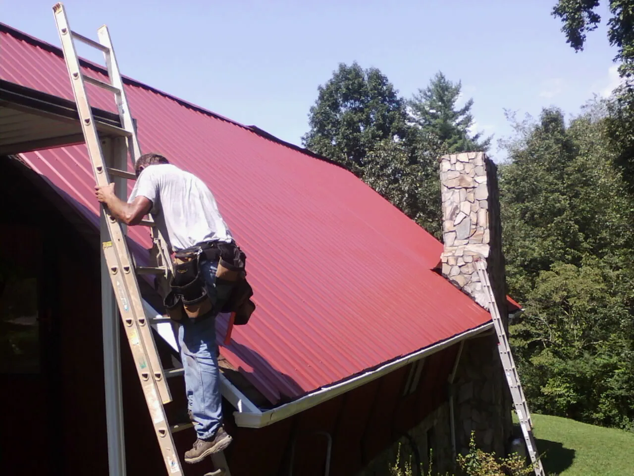 Roofer on a ladder beside a red metal roof and stone chimney — Soffit & Fascia Repair by Roofing Brothers in South Haven